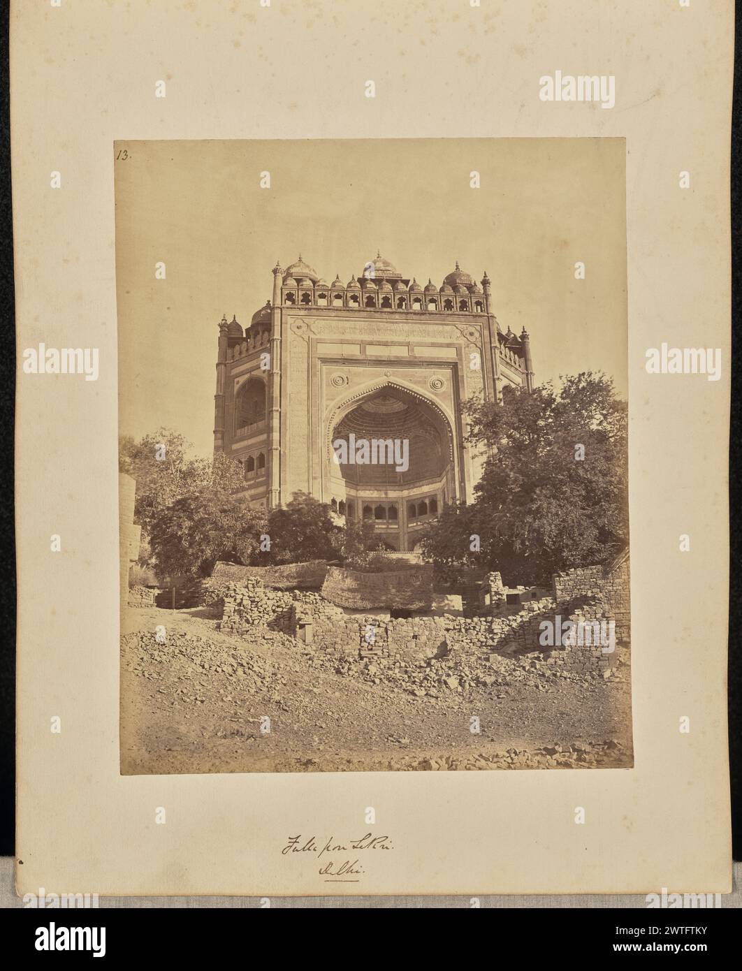 Futtepore Sekri. Delhi. John Edward Saché, Fotograf (preußisch oder britisch, geb. Preußen, 1824–1882) um 1868 Ansicht der Buland Darwaza, ein monumentales Tor mit einem großen Iwan oder einem dreiseitigen gewölbten Saal, gekrönt von einer Reihe von Chattris oder dekorativen Kuppelpavillons. Zwei Hütten und die Ruinen von Ziegelmauern sind im Vordergrund unter dem Tor zu sehen. (Recto, bedruckt) oben links, in brauner Tinte: '13.'; unten rechts, im negativ beschriftet: '360'; (Recto, Mount) unten in der Mitte, in brauner Tinte: 'Futtepore Sekri. / Delhi. [Unterstrichen]'; (Rückseite, Halterung) unten rechts, in Bleistift: 'A48.13 (Saché) [ Stockfoto