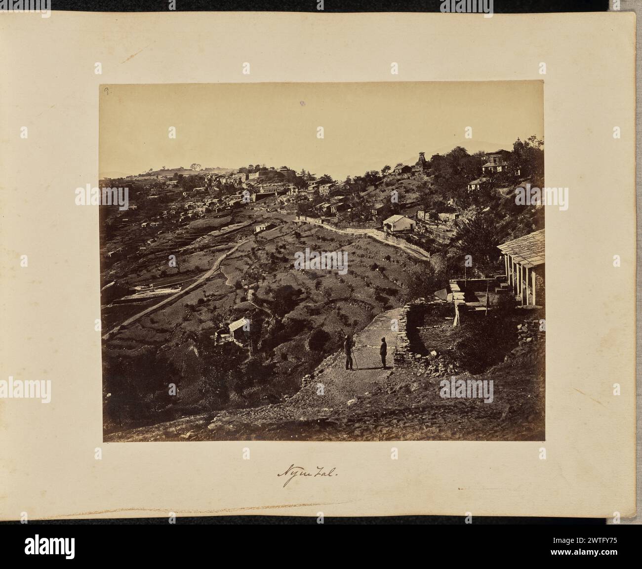 Nynee Tal. John Edward Saché, Fotograf (preußisch oder britisch, geb. Preußen, 1824–1882) etwa 1869 zwei Männer stehen auf einem unbefestigten Weg vor einem Hanghaus. Einer der Männer hält einen Stock. Hinter ihnen befindet sich die Stadt Almora am Hügel mit ihrem terrassenförmig angelegten Tal und dem Nanda Devi Tempel, der rechts oben sichtbar ist. Die Chihara des Tempels, oder glockenförmige Kuppel, die typischerweise auf Hindu-Tempeln zu finden ist, ist mit einer galerieartigen Struktur gekrönt. (Recto, Druck) oben links, in brauner Tinte: '9.'; unten Mitte, im negativ beschriftet: '484'; (Recto, Mount) unten Mitte, in brauner Tinte: 'Nynee Tal. [ Stockfoto Nynee Tal. John Edward Saché, Fotograf (preußisch oder britisch, geb. Preußen, 1824–1882) etwa 1869 zwei Männer stehen auf einem unbefestigten Weg vor einem Hanghaus. Einer der Männer hält einen Stock. Hinter ihnen befindet sich die Stadt Almora am Hügel mit ihrem terrassenförmig angelegten Tal und dem Nanda Devi Tempel, der rechts oben sichtbar ist. Die Chihara des Tempels, oder glockenförmige Kuppel, die typischerweise auf Hindu-Tempeln zu finden ist, ist mit einer galerieartigen Struktur gekrönt. (Recto, Druck) oben links, in brauner Tinte: '9.'; unten Mitte, im negativ beschriftet: '484'; (Recto, Mount) unten Mitte, in brauner Tinte: 'Nynee Tal. [ Stockfoto
