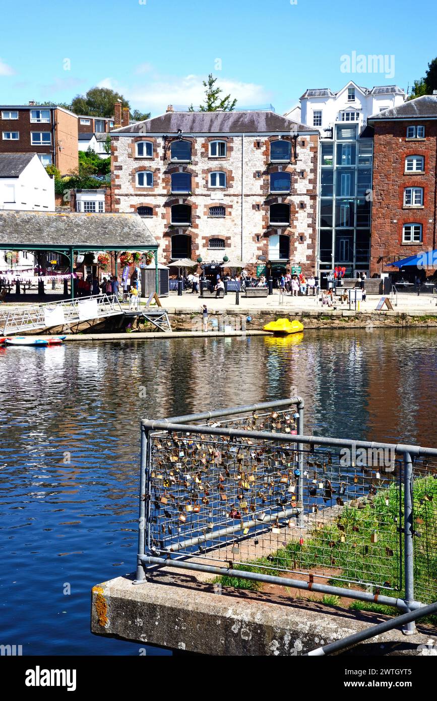 Blick über den Fluss exe zu Geschäften und Restaurants entlang des East Quay mit Lovelocks auf einem Zaun im Vordergrund, Exeter, Devon, Großbritannien, Europa. Stockfoto