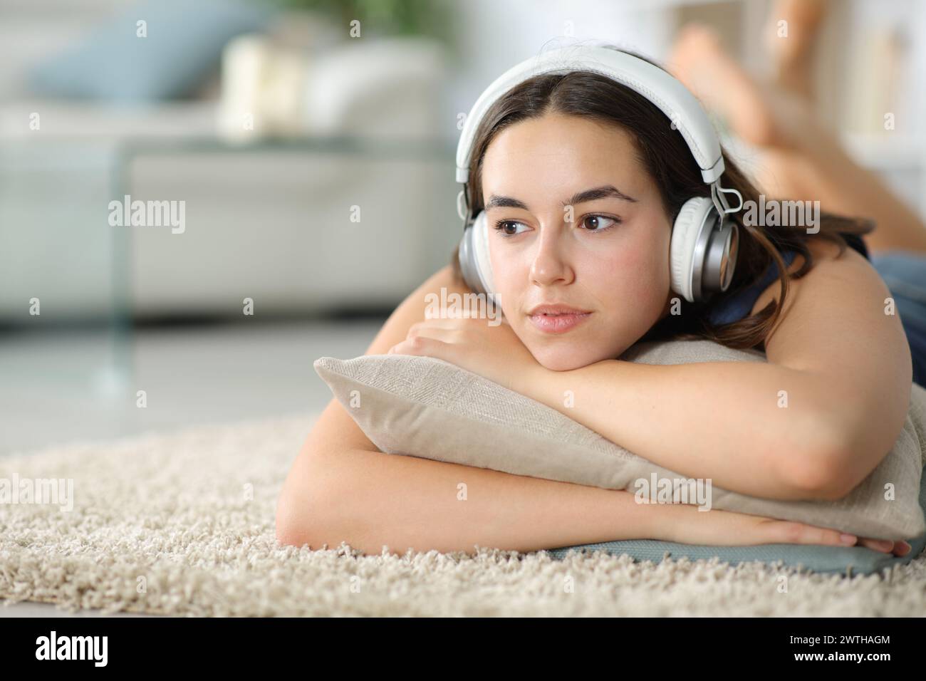 Nachdenkliche Frau mit Kopfhörern, die Musik hört, die auf einem Teppich liegt Stockfoto