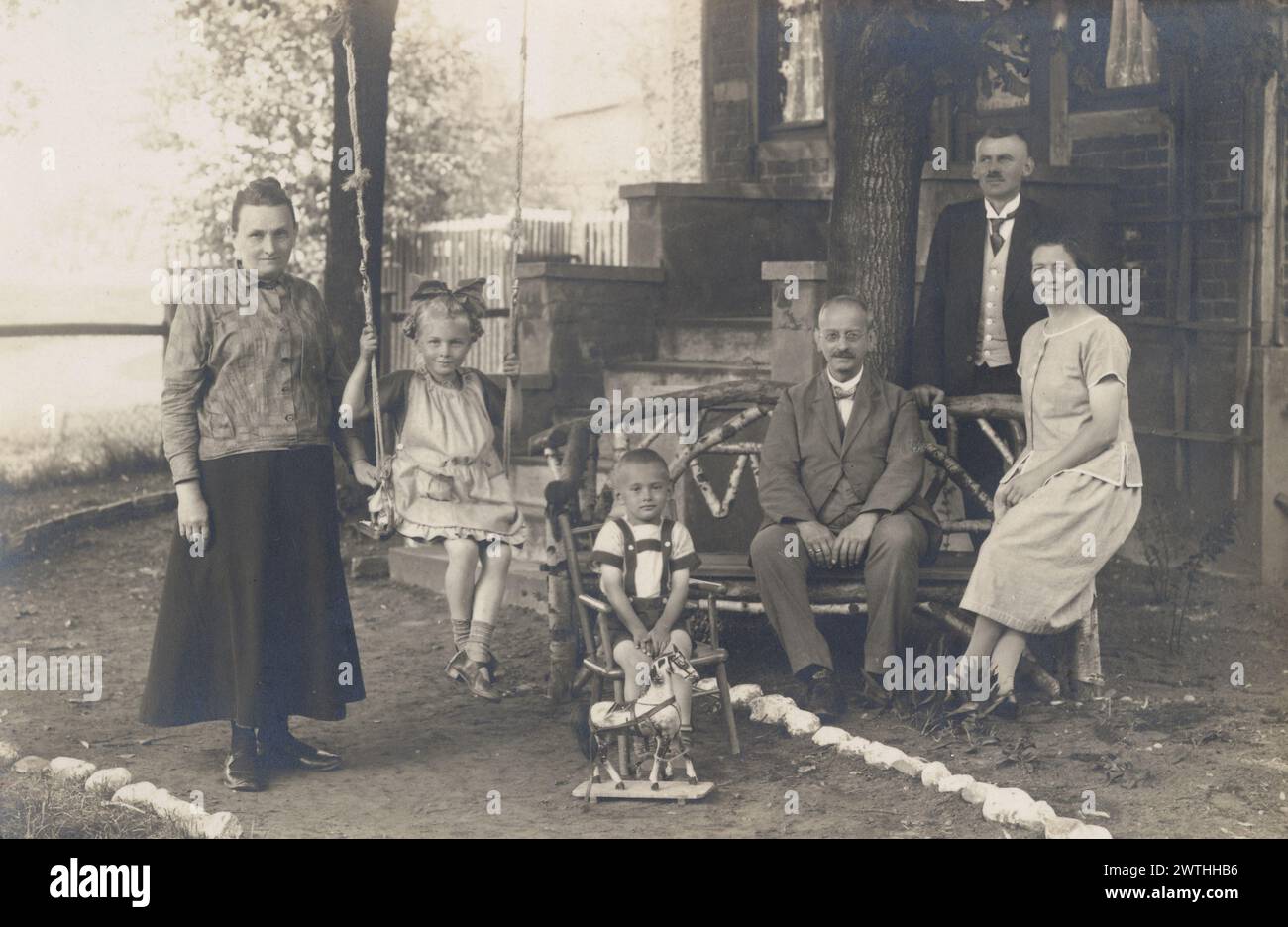 Familie auf Veranda vor Haus C1910. In der Stadt Neuhaldensleben. Eltern und Großeltern, mit kleinen Kindern, einer auf einer Schaukel, der andere mit Holzspielpferd. Sehr detaillierte Moden. Sozialgeschichte. Foto Stockfoto