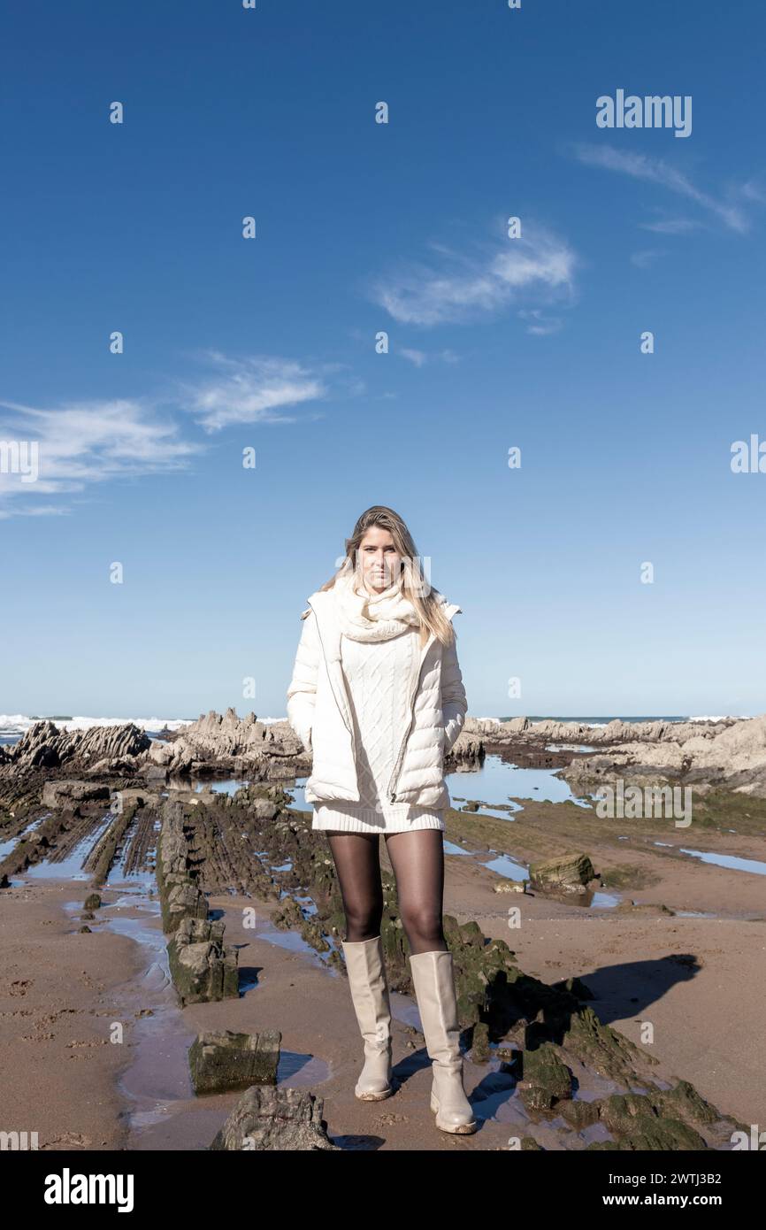 Frau steht am Strand mit weißem Kleid und Stiefeln. Der Himmel ist blau und im Hintergrund sind Wolken Stockfoto