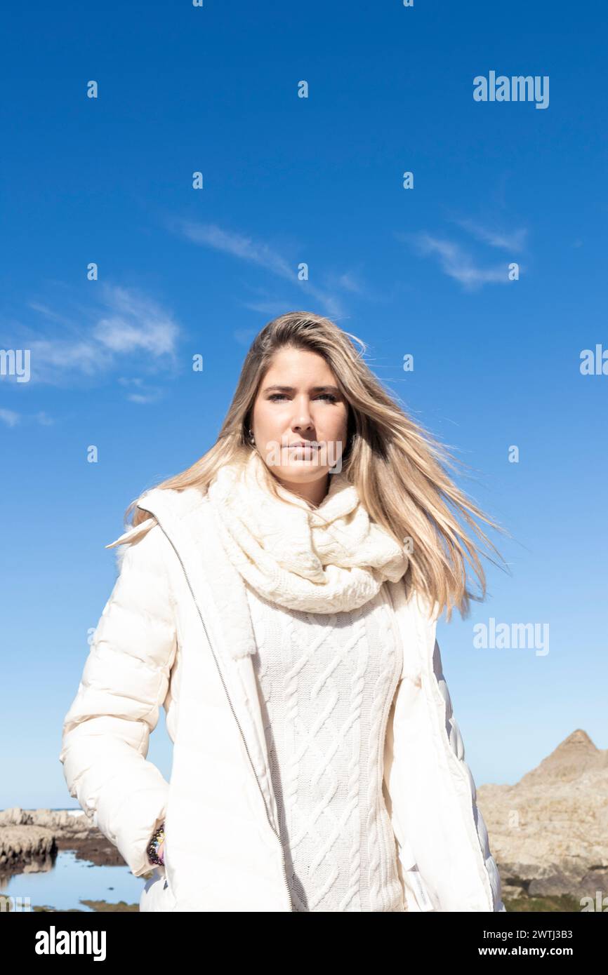 Eine Frau mit weißer Jacke und Schal steht vor einem blauen Himmel. Sie sieht selbstbewusst aus, mit ihren Haaren, die im Wind wehen Stockfoto