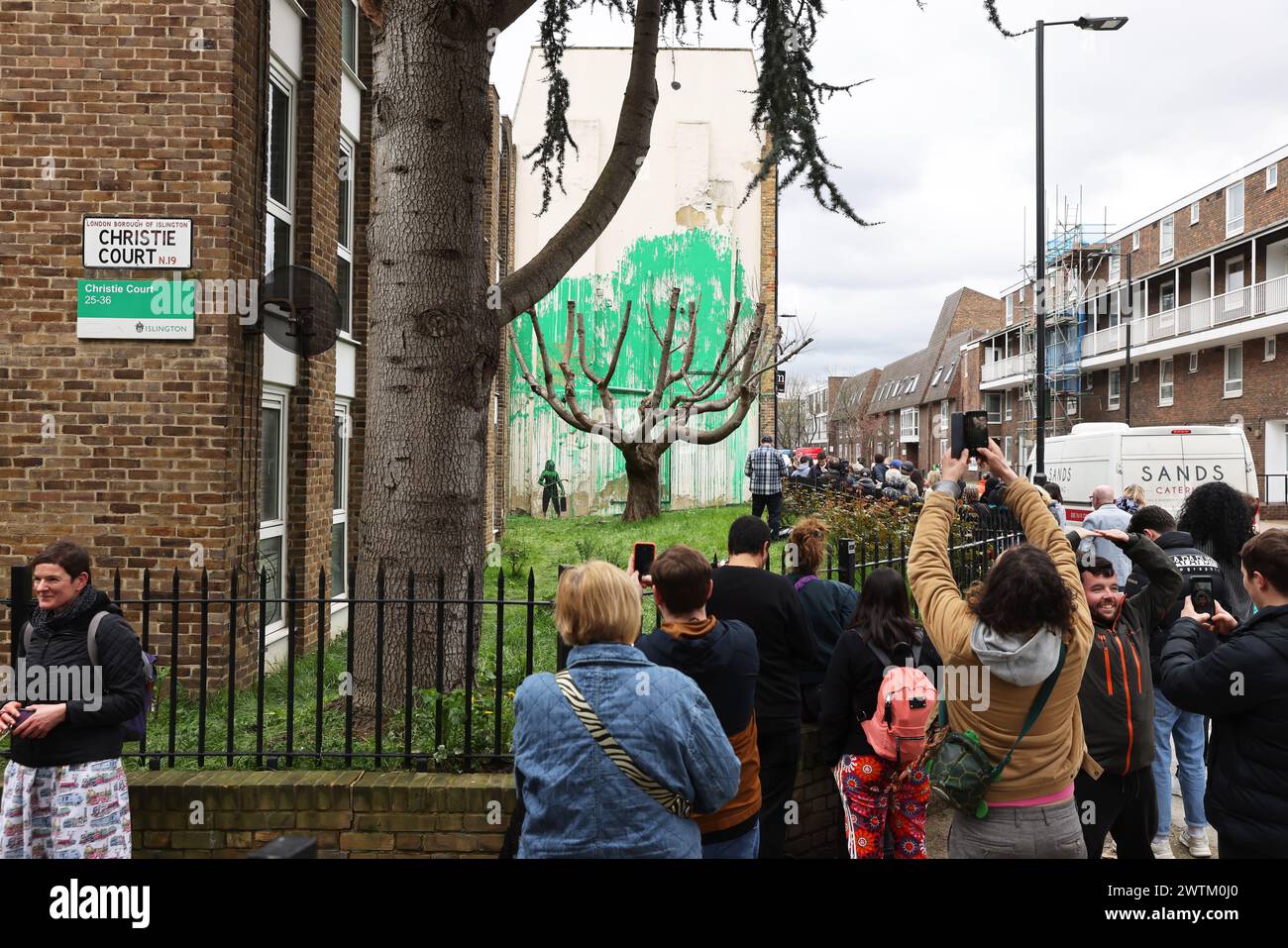 London, Großbritannien, 18. März 2024. Besucher haben sich in der Hornsey Street in der Nähe des Finsbury Park im Norden Londons versammelt, um Banksys neues Wandgemälde zu sehen. Grüne Farbe wurde auf eine Wand hinter dem Baum gesprüht, um die Illusion von Laub zu erwecken. Eine Person hält einen Druckschlauch daneben. Die grüne Farbe scheint der Beschilderung des Islington Council zu ähneln, und es gibt auch eine mögliche Verbindung zum St. Patrick's Day gestern. Kredit : Monica Wells/Alamy Live News Stockfoto