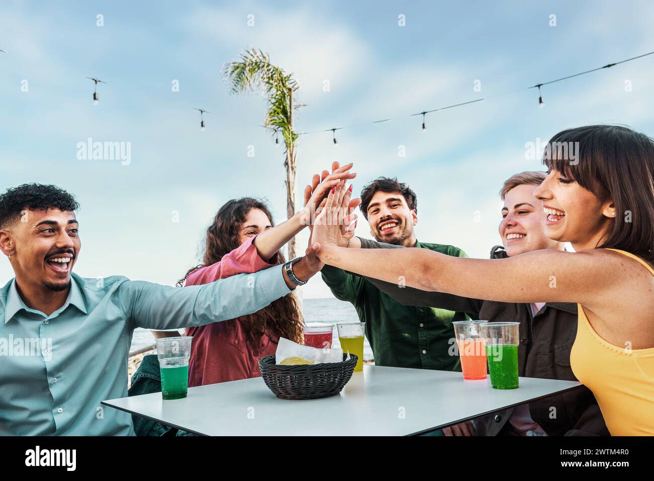 Freunde vereinen sich in einem High-Five - Lachen und bunte Getränke am Meer - den Geist der Zweisamkeit unter freiem Himmel Stockfoto