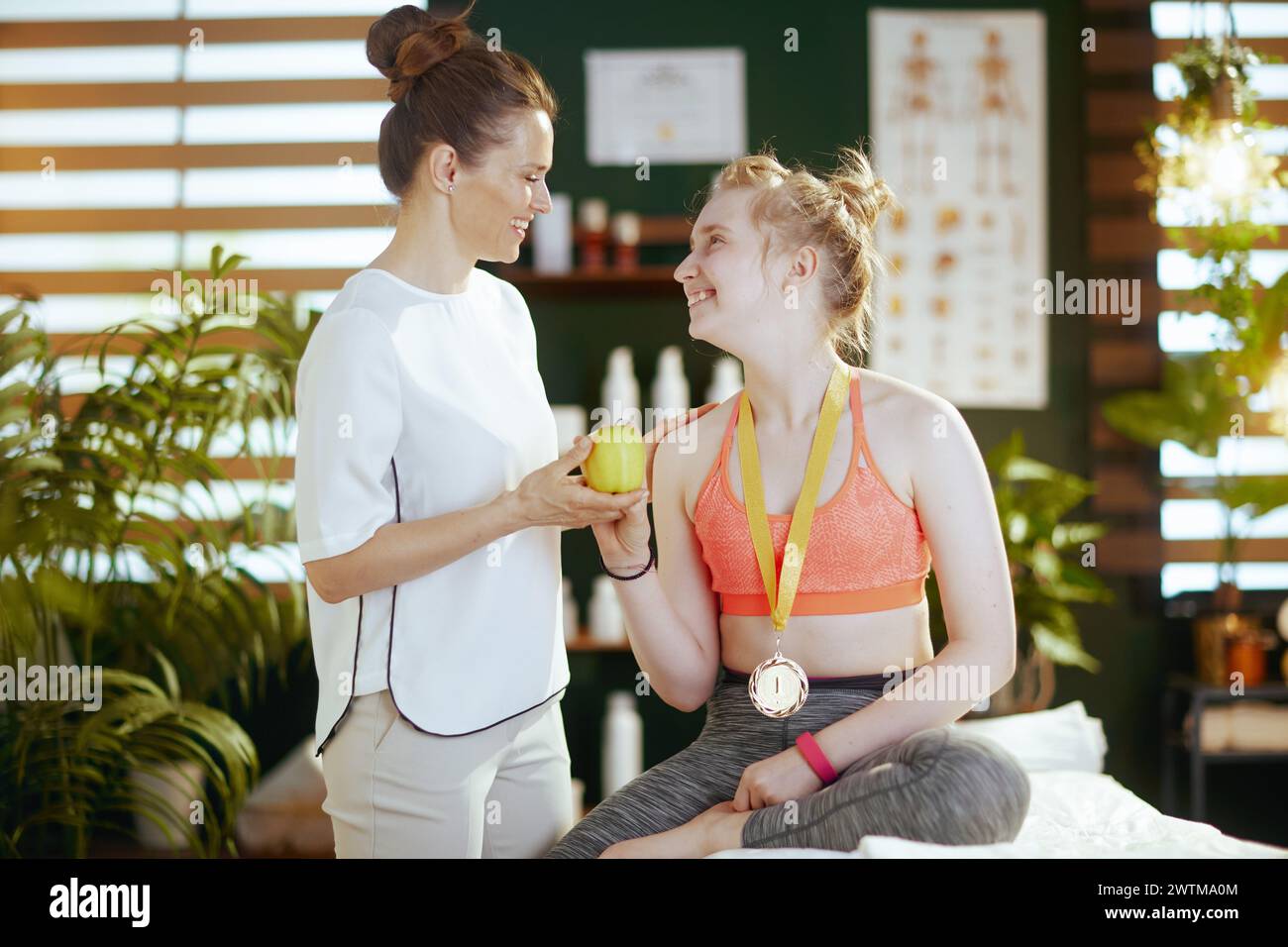 Gesundheitszeit. Lächelnde Masseurin in Massageschrank mit Teenager-Klientin, einem Apfel und einer goldenen Medaille. Stockfoto