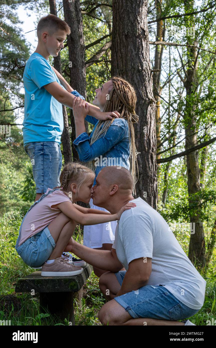In einem üppigen Waldpark teilt eine Familie einen Moment der Zuneigung und Verbundenheit, wobei Eltern ihre Liebe zu ihrem Sohn und ihrer Tochter zeigen und die Eltern verkörpern Stockfoto