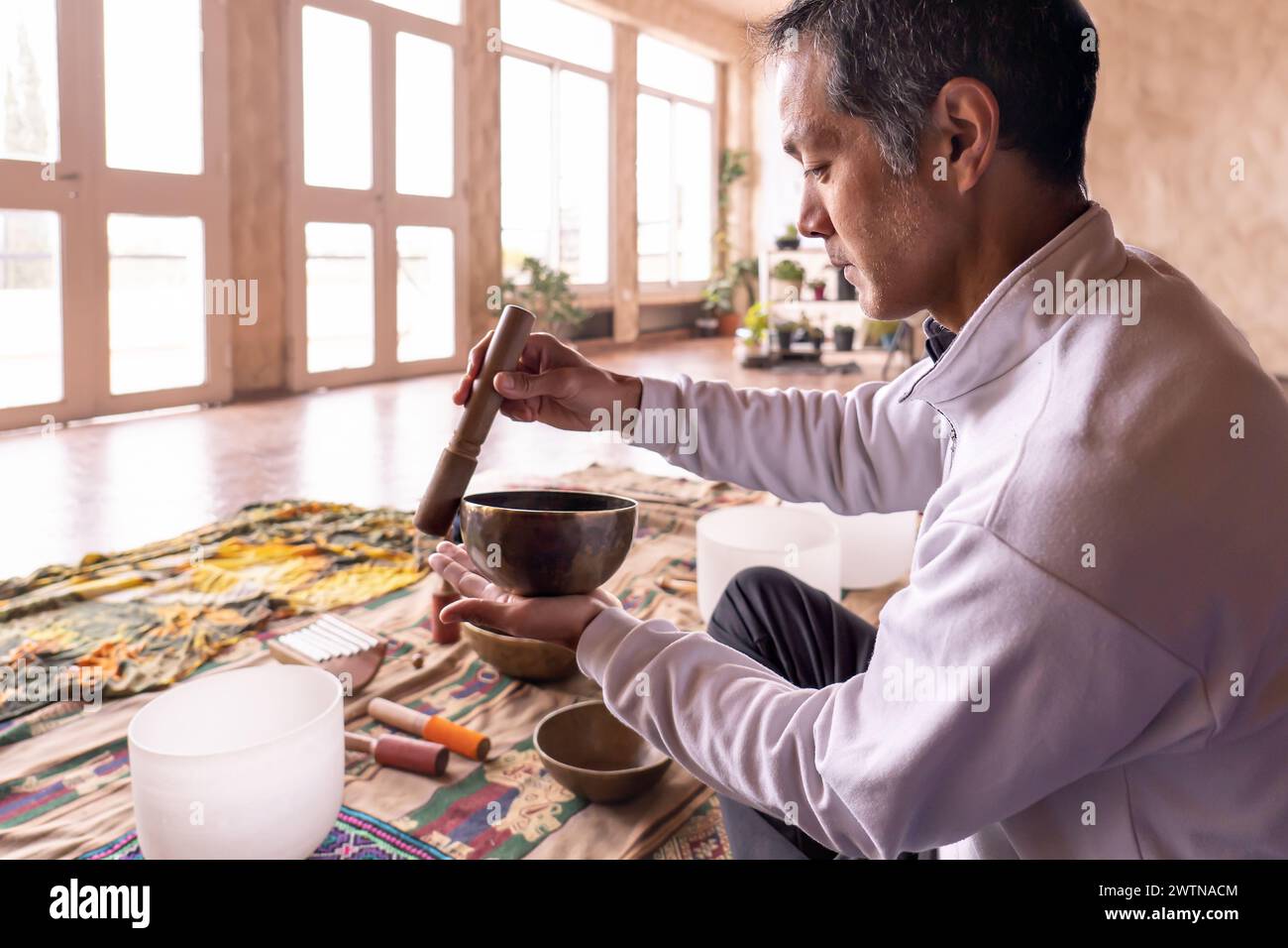Ein asiatischer Mann mittleren Alters sitzt im Profil und konzentriert sich auf sanfte Vibrationen einer Messingschale in einem sanft beleuchteten Raum. Antike Praxis der Klangmeditation in Stockfoto