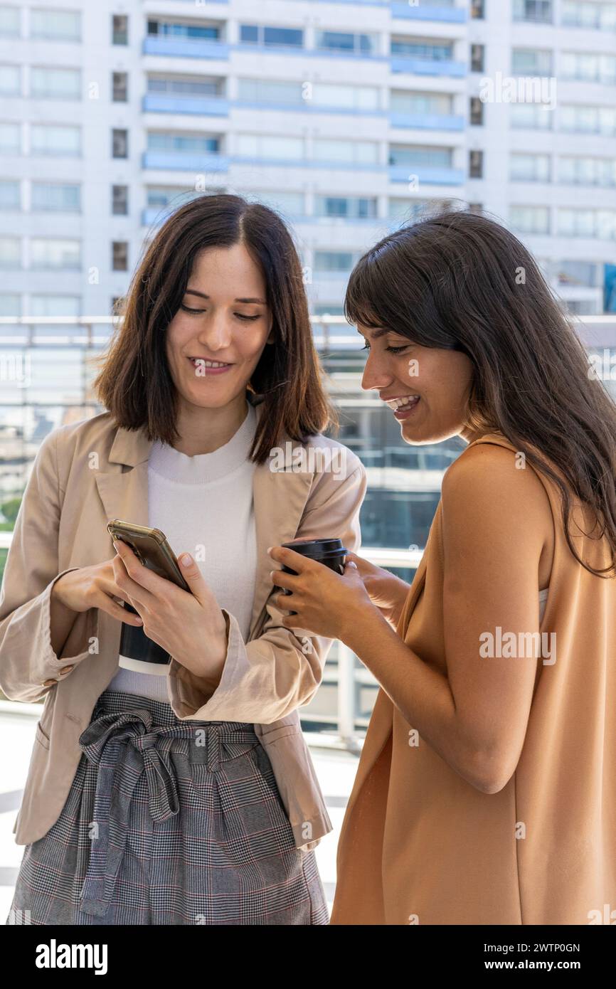 Zwei Frauen sind draußen. Eine zeigt etwas auf ihrem Handy, während die andere eine Tasse hält. Stockfoto