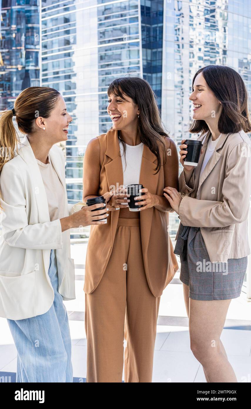 Drei Geschäftsfrauen stehen draußen vor einem Gebäude, genießen ihre Büropause, lächeln und halten Tassen Kaffee. Das Konzept des Camaraders Stockfoto