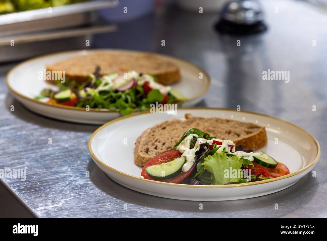 Dieses Bild zeigt zwei Teller mit frisch zubereiteten Salaten in einem Restaurant, die jeweils von einer Scheibe Vollkornbrot begleitet werden. Die Salate sind gut zusammengesetzt und umfassen eine Auswahl an Gemüse, Tomaten, Gurken und Zwiebeln, leicht gekleidet und bereit zum Servieren. Die Küchenzeile bietet eine saubere und professionelle Kulisse, die den Fokus auf die Präsentation von Speisen unterstreicht. Frisch zubereitete Salate auf Restaurantplatten. Hochwertige Fotos Stockfoto