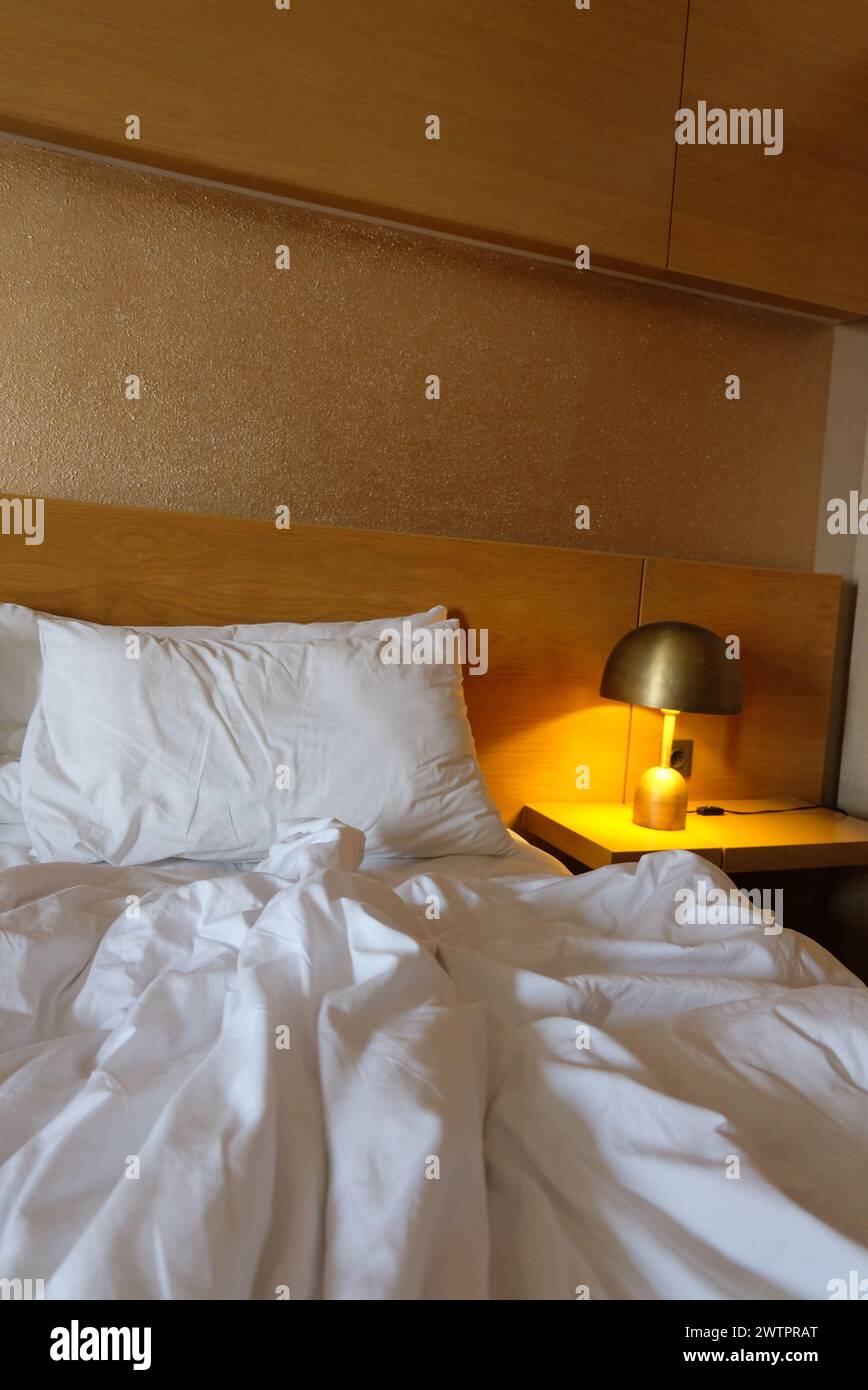 Selektiv fokussiert auf ein unordentliches Schlafzimmer mit Lampe in der Nähe des Bettes. Die Bettwäsche und die Kissen sind zerknittert. Schmutziges Schlafzimmer, weißes Kissen mit Decke, die ungemacht auf dem Bett lag. Stockfoto Selektiv fokussiert auf ein unordentliches Schlafzimmer mit Lampe in der Nähe des Bettes. Die Bettwäsche und die Kissen sind zerknittert. Schmutziges Schlafzimmer, weißes Kissen mit Decke, die ungemacht auf dem Bett lag. Stockfoto