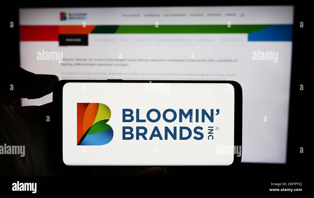 Person, die ein Mobiltelefon mit dem Logo der US-Restaurantholding Bloomin' Brands Inc. Vor der Webseite des Unternehmens hält. Schwerpunkt auf dem Display des Telefons. Stockfoto