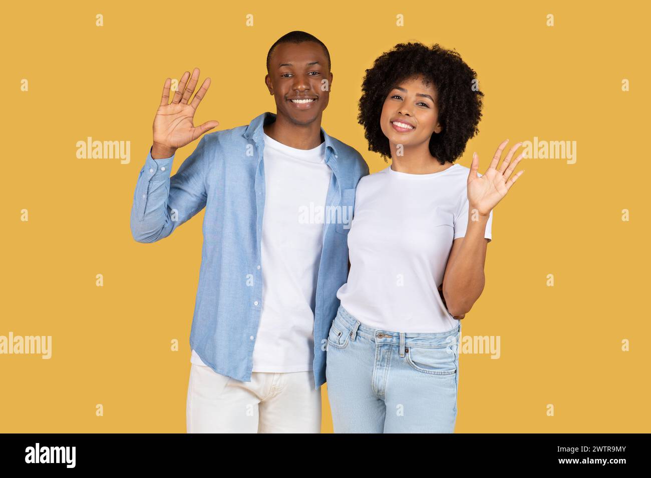 Glückliches afroamerikanisches Paar, das die Hände mit hellem Lächeln zur Kamera winkt, lässig gekleidet Stockfoto