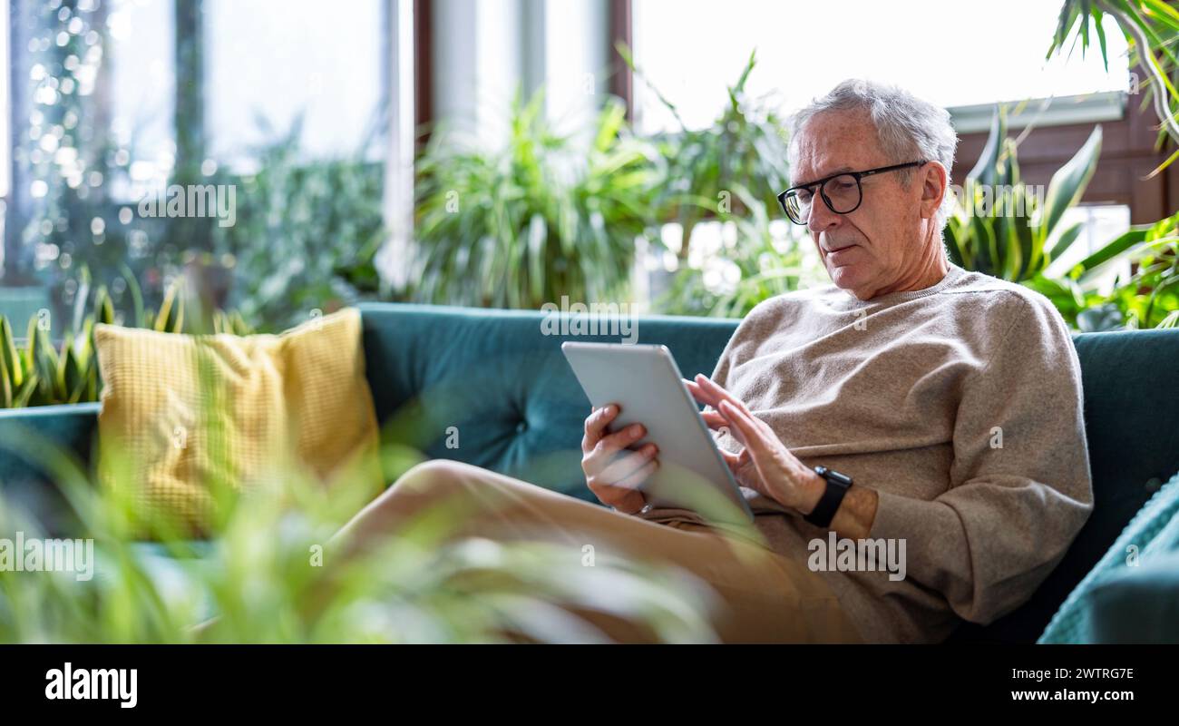 Seniorenmann, der ein digitales Tablet verwendet, während er zu Hause im Wohnzimmer auf dem Sofa sitzt Stockfoto