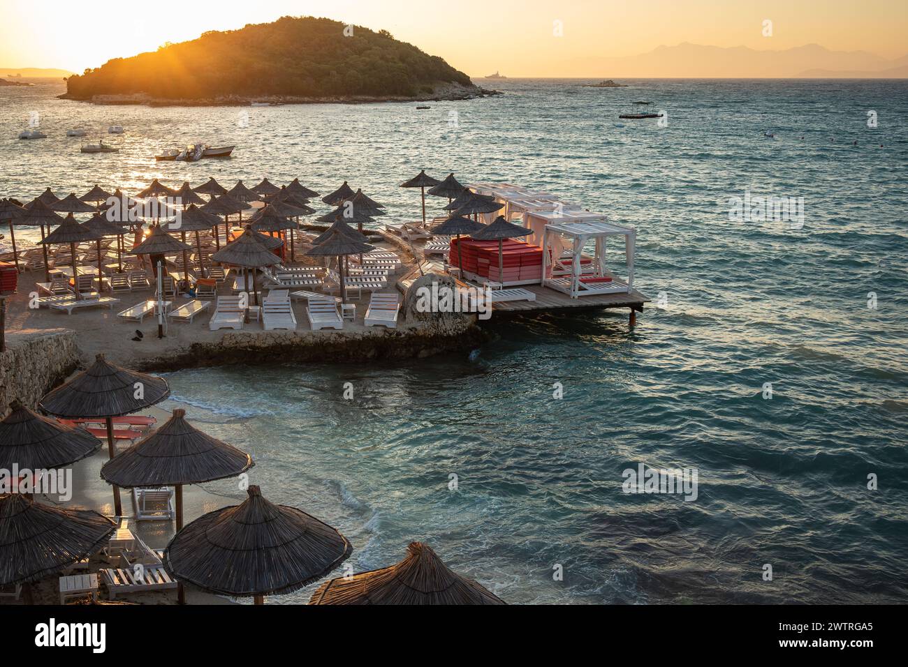 Strandlandschaft von Ksamil Beach mit seiner Insel bei Sonnenuntergang. Sommerabend mit Ionischem Meer und Sonnenliegen mit Sonnenschirmen an der albanischen Riviera. Stockfoto