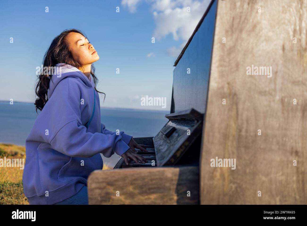 Eine attraktive Asiatin hat die Augen geschlossen und spielt draußen Klavier. Ein charmantes koreanisches Mädchen spielt gern ein Musikinstrument in der Natur. Stockfoto