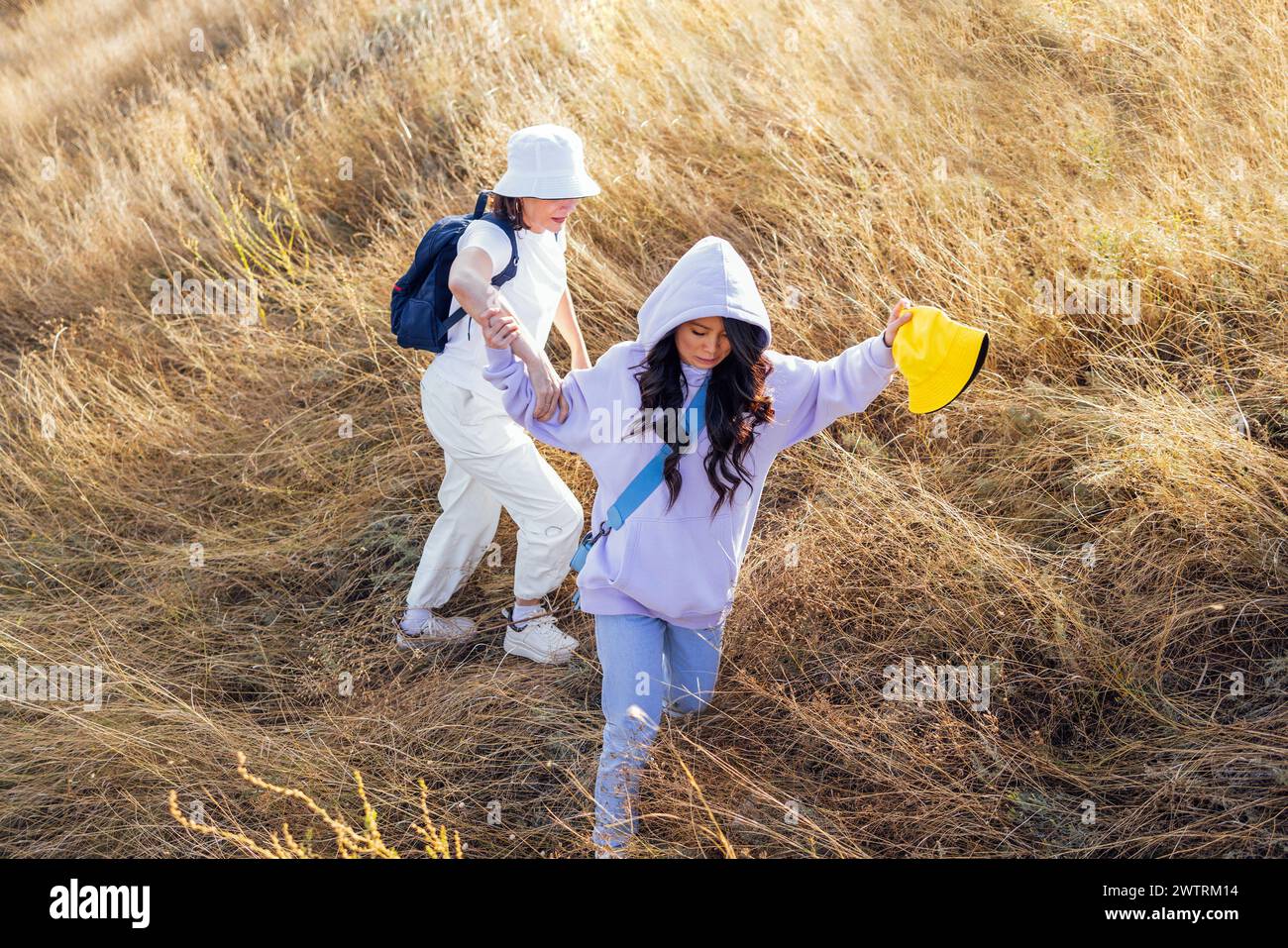 Zwei attraktive, multiethnische Freunde in lässiger Kleidung und Sommerhüten laufen durch das hohe, trockene Gras. Mädchen verschiedener Rassen gehen wandern. Jung W Stockfoto