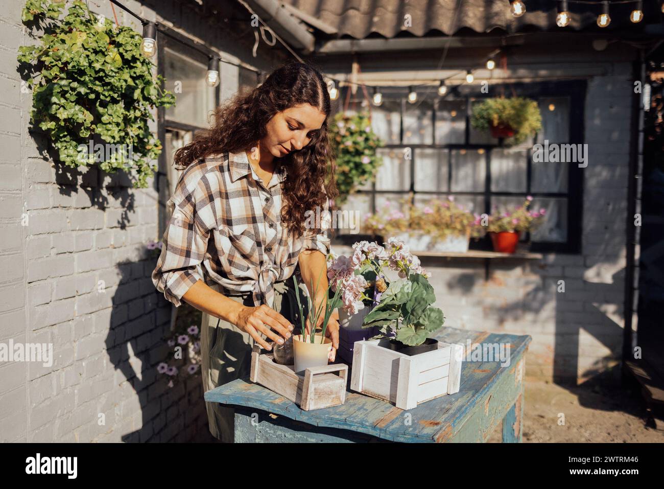 Eine junge charmante Frau in lässiger Kleidung pflanzt Blumen auf einem Tisch draußen. Ein süßes Mädchen mit lockigen Haaren pflanzt grüne Pflanzen in einen Topf in t Stockfoto