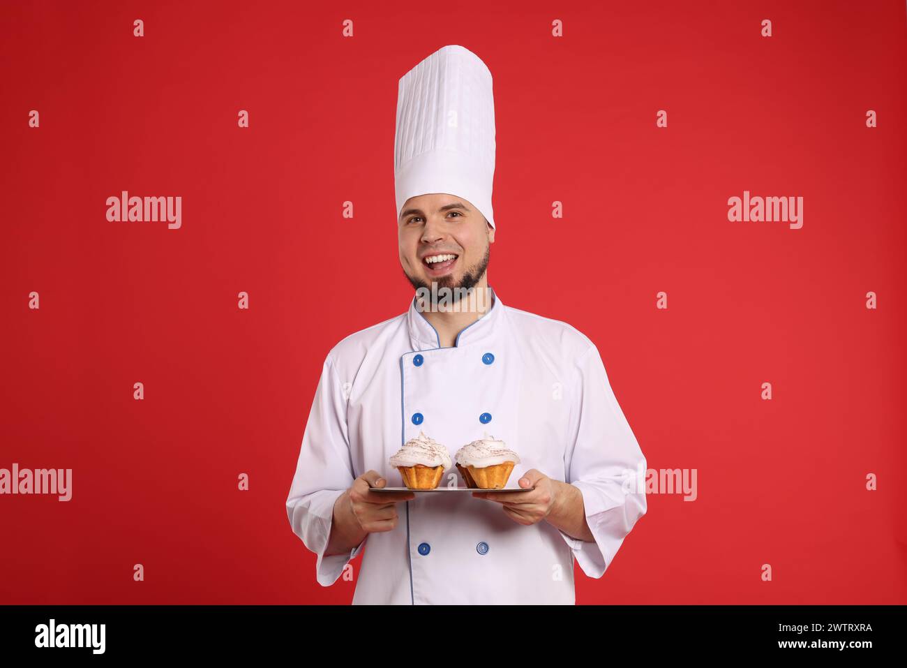 Glücklicher professioneller Konditor in Uniform mit köstlichen Cupcakes auf rotem Hintergrund Stockfoto