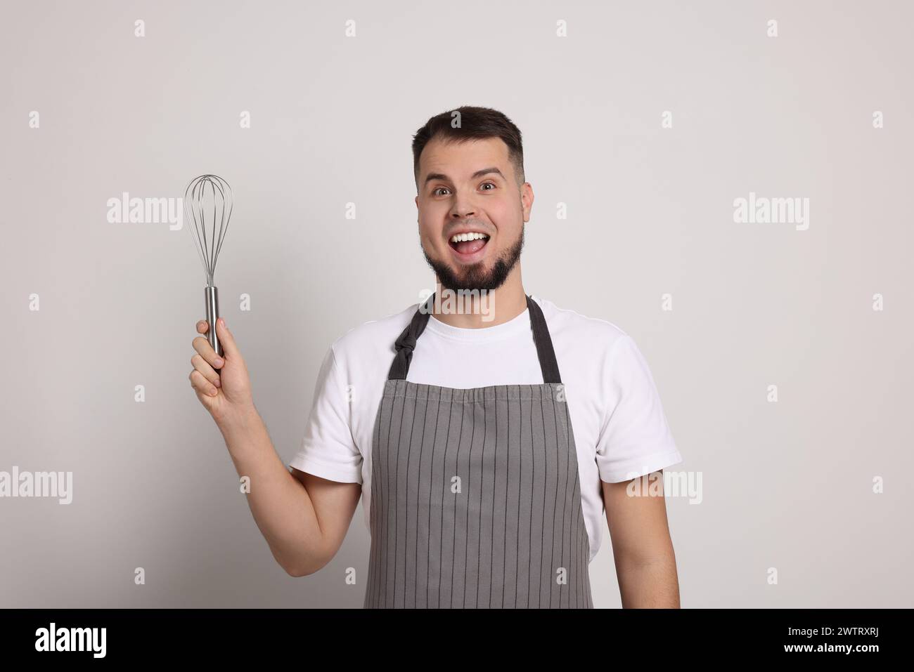 Begeisterte professionelle Konditorin in Schürze mit Schneebesen auf hellgrauem Hintergrund Stockfoto