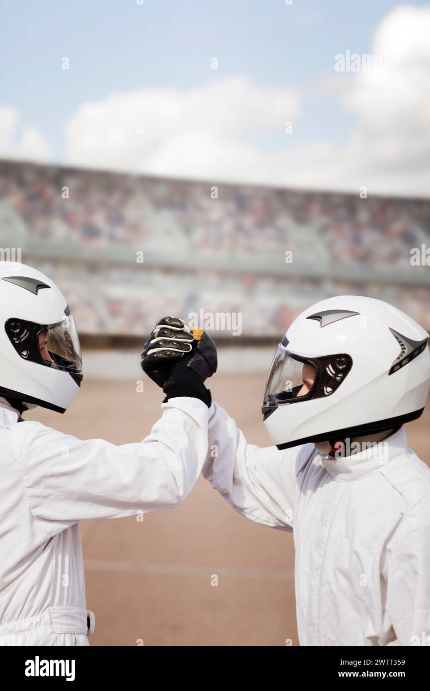 Zwei Rennfahrer in weißen Helmen teilen sich einen Faustschlag vor einer dynamischen Rennstrecke. Stockfoto