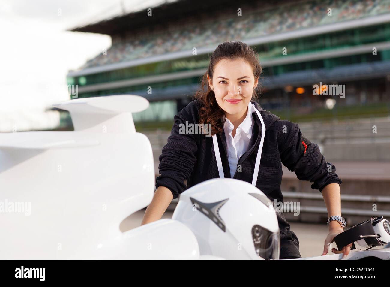 Selbstbewusste Frau mit einem hellen Lächeln, die sich auf ein Rennmotorrad auf einer Rennstrecke stützt. Stockfoto