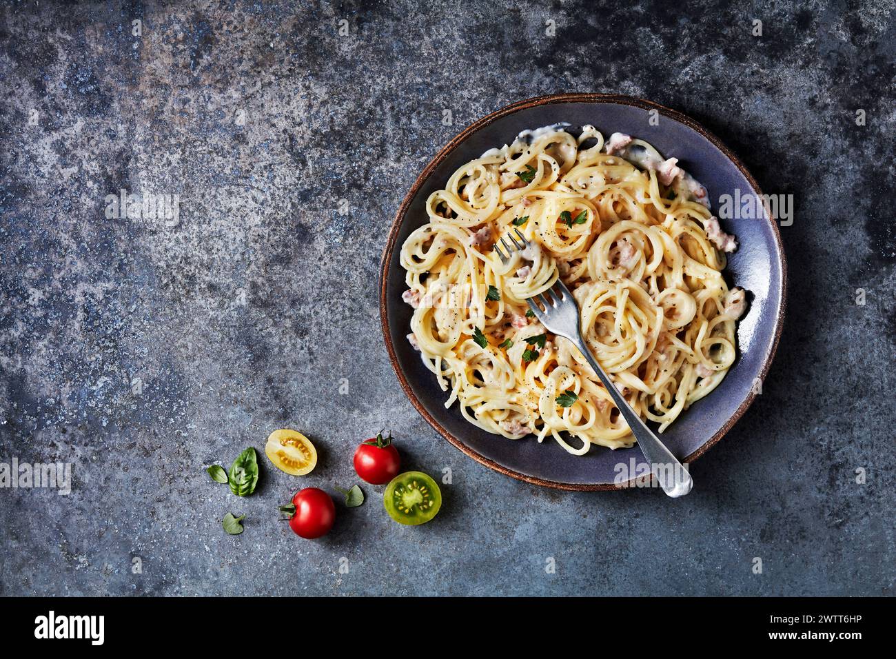 Eine köstliche Schüssel cremiger Pasta auf strukturierter grauer Oberfläche, begleitet von frischen Tomaten und Basilikumblättern. Stockfoto