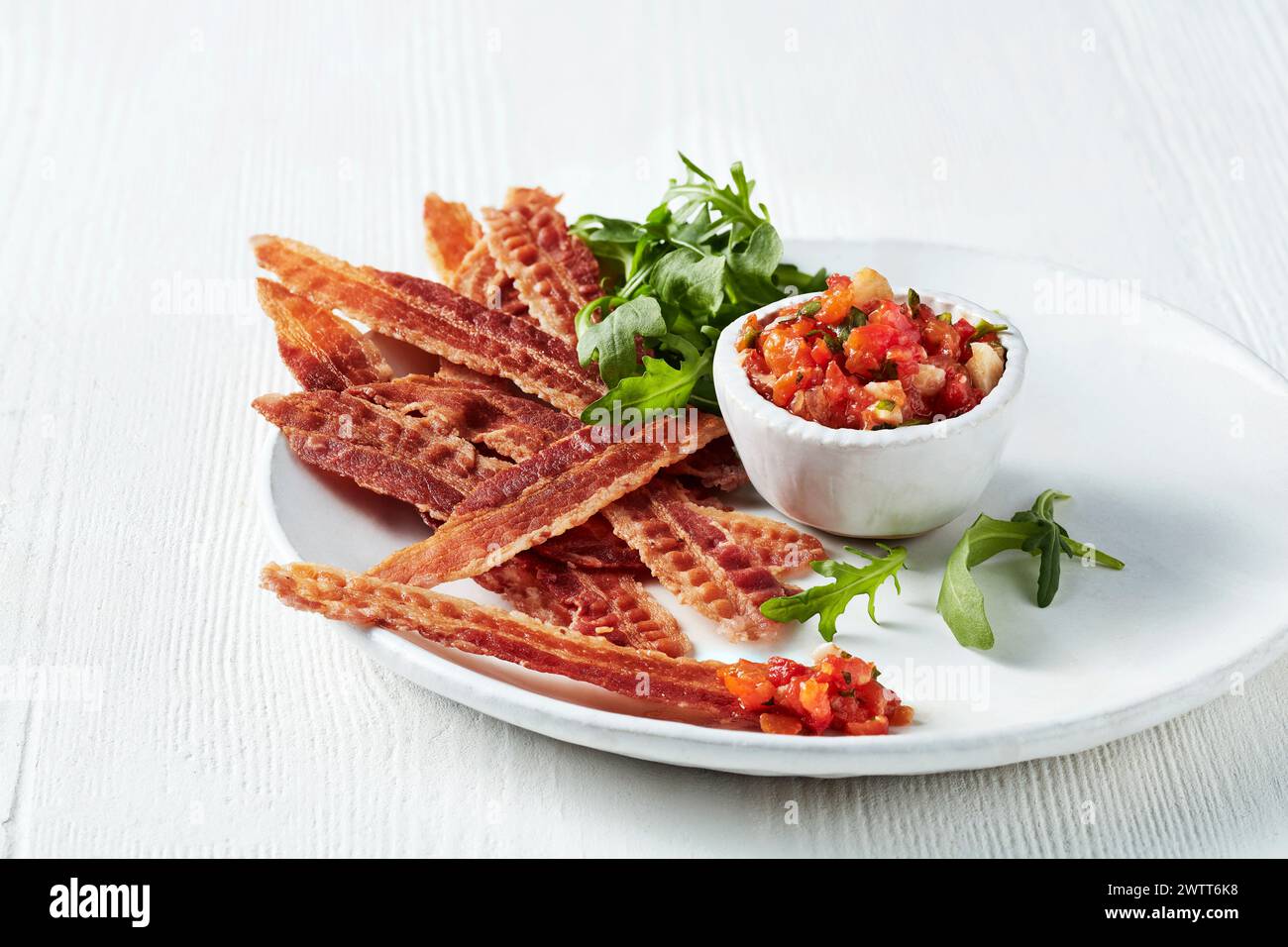 Knusprige Speckstreifen, serviert mit einer Seite frischer Salsa und garniert mit Rucola auf einem weißen Teller. Stockfoto
