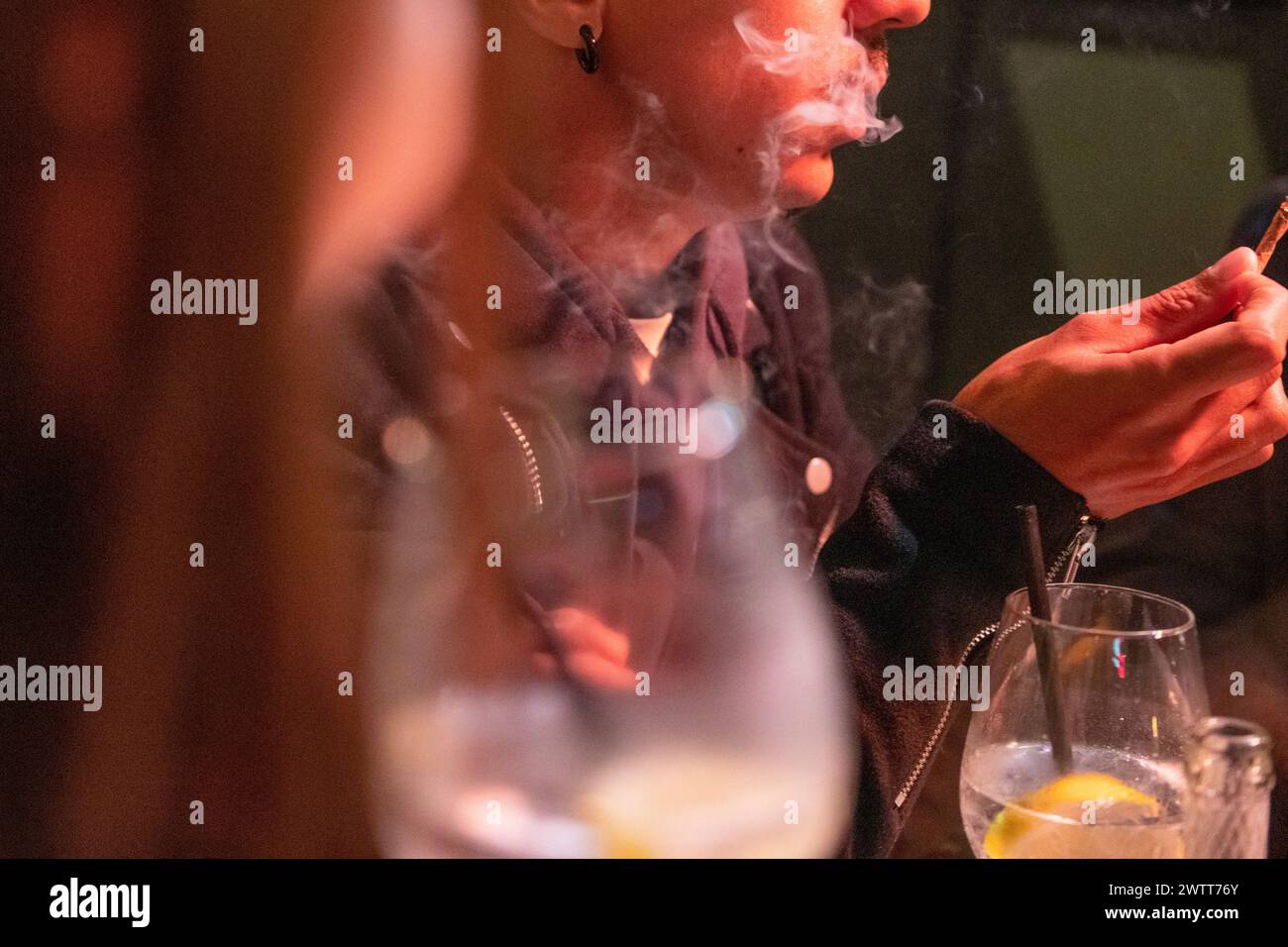 Eine Person atmet Rauch aus, während sie ihr Smartphone an einem schwach beleuchteten Tisch durchsucht. Stockfoto