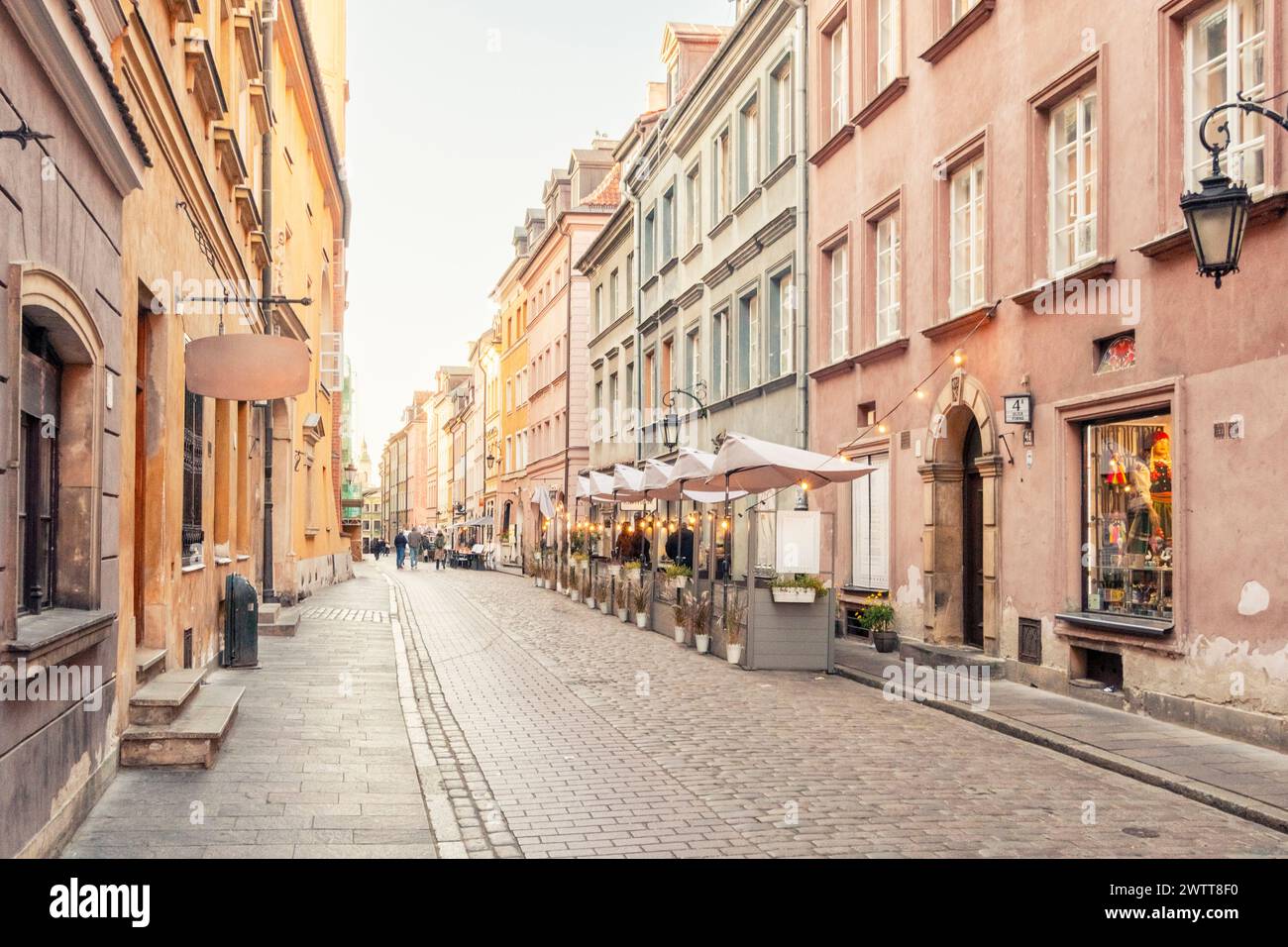 Die gepflasterte Straße, die bei Sonnenuntergang in ein sanftes, warmes Licht getaucht ist, gesäumt von malerischen Geschäften und Restaurants in einer historischen europäischen Stadt. Stockfoto