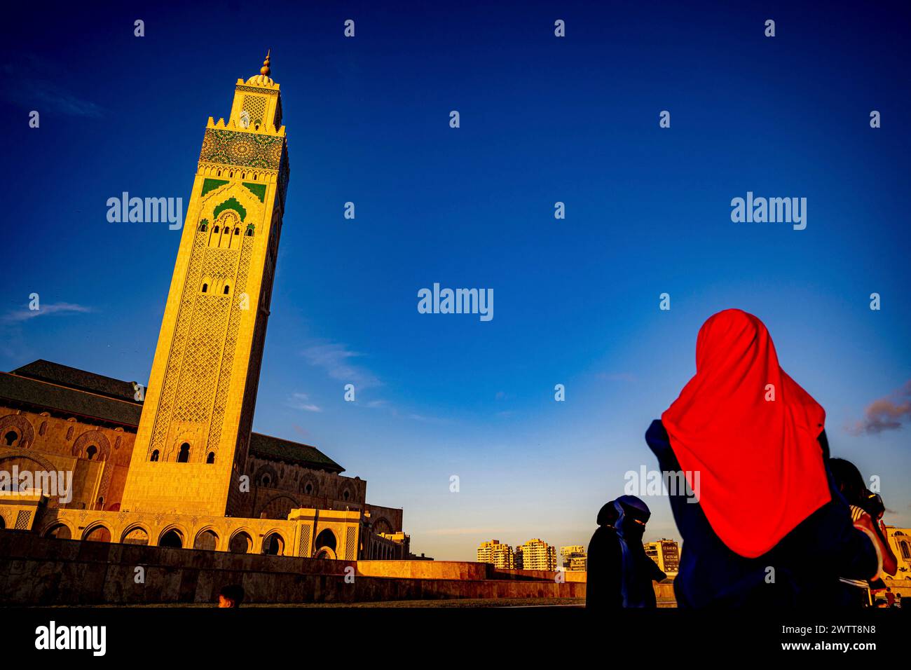 Eine Person in einem leuchtend roten Kopftuch blickt bei Sonnenuntergang auf einen majestätischen Moschee-Turm vor einem tiefblauen Himmel. Stockfoto