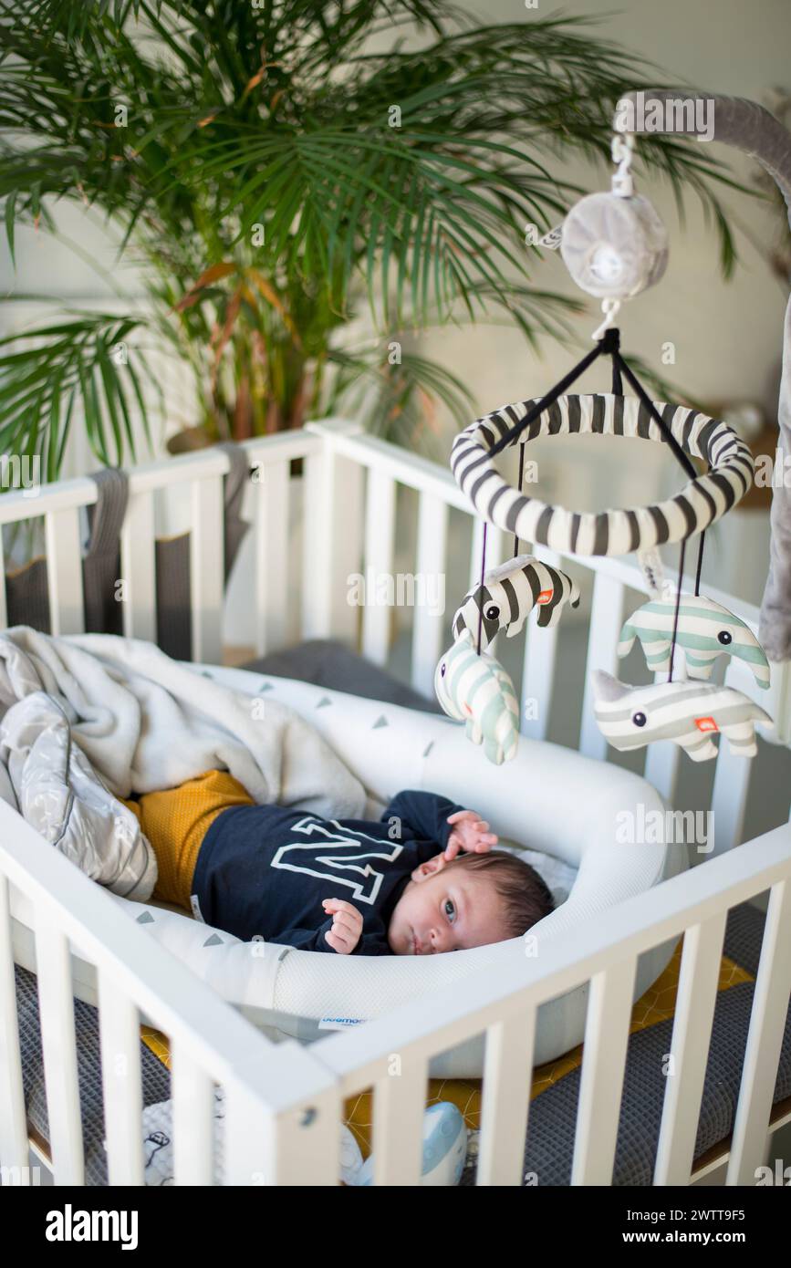Das Baby blickt neugierig in einem weißen Kinderbett mit einem Mobile darüber. Stockfoto