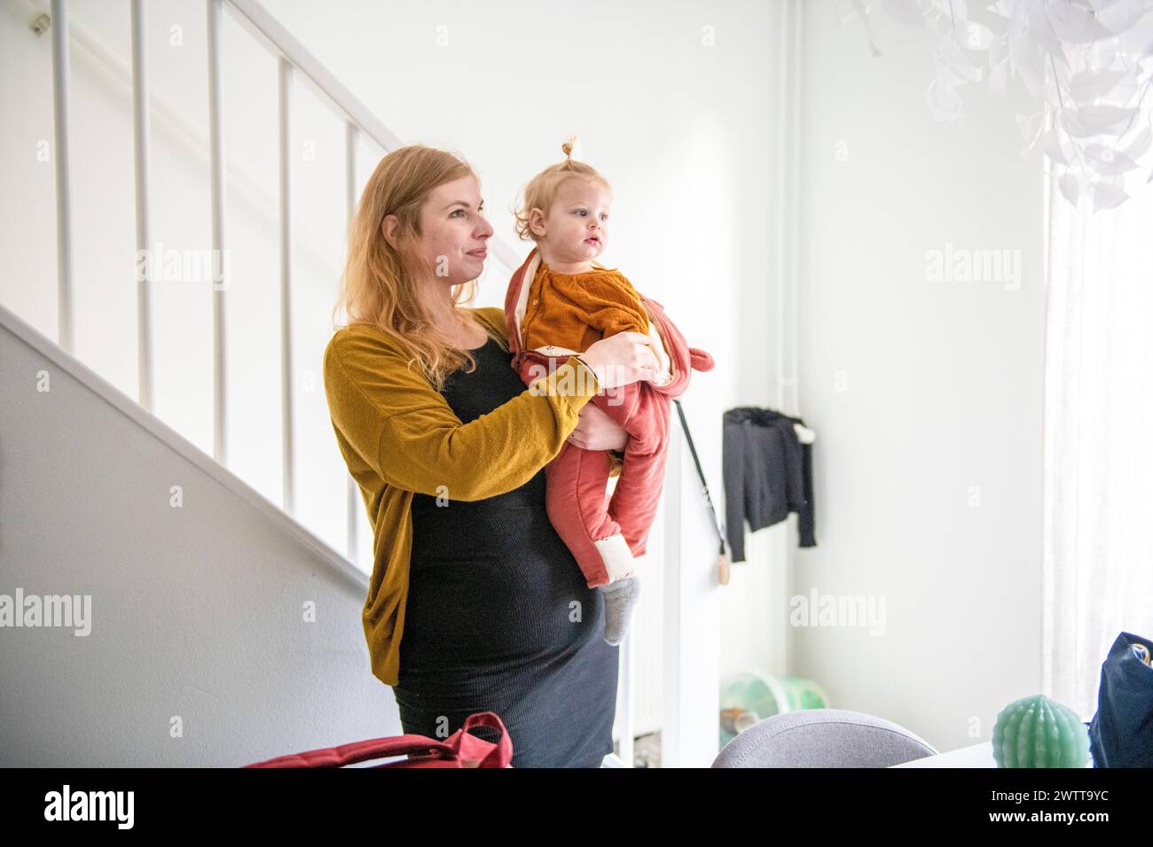 Mutter hält ihr kleines Kind, während sie an der Treppe steht, und blickt mit sanfter Neugier nach vorne. Stockfoto