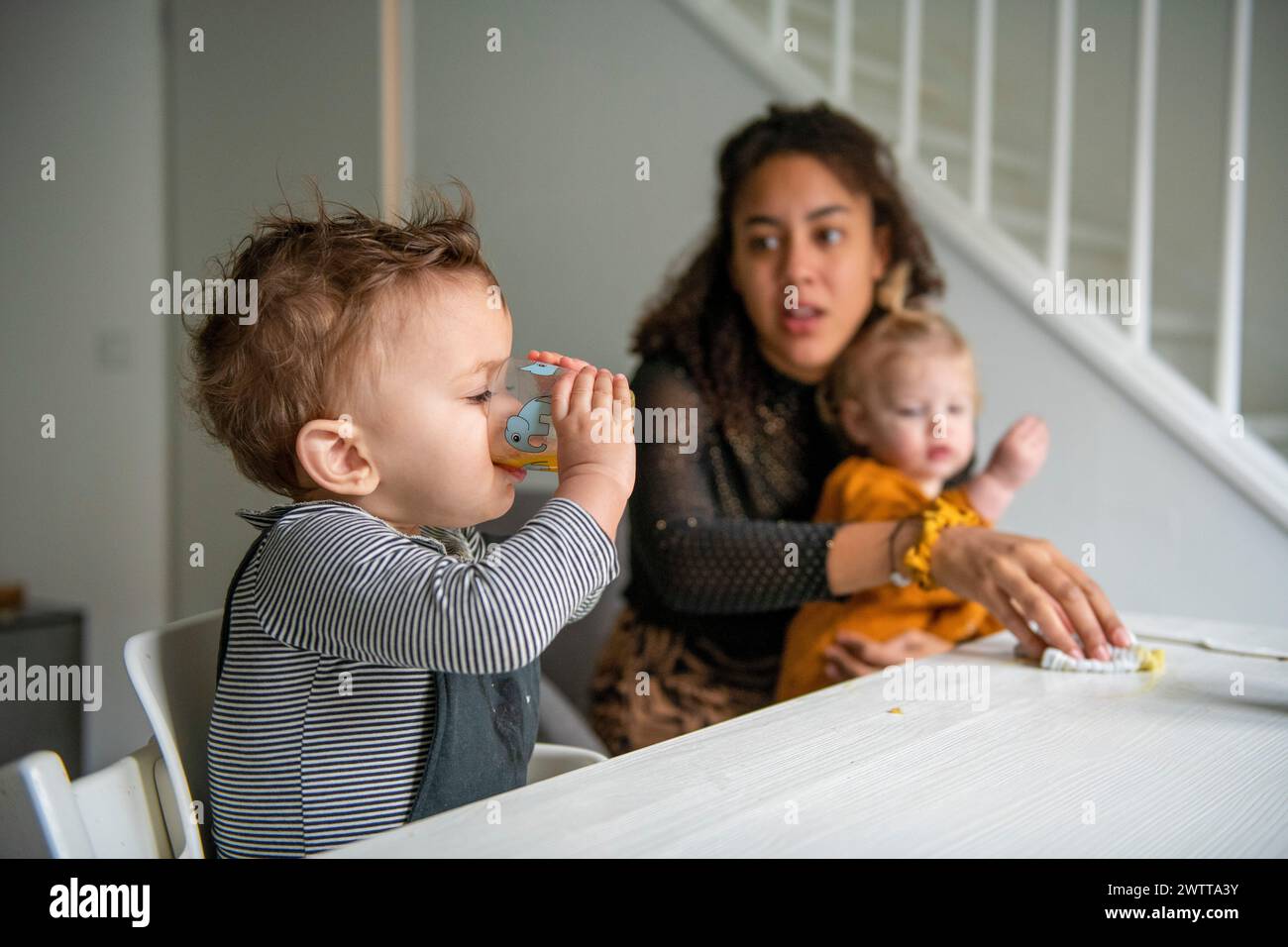 Ein Kleinkind schlürft am Esstisch aus einer Tasse, während Mom über sie wacht. Stockfoto