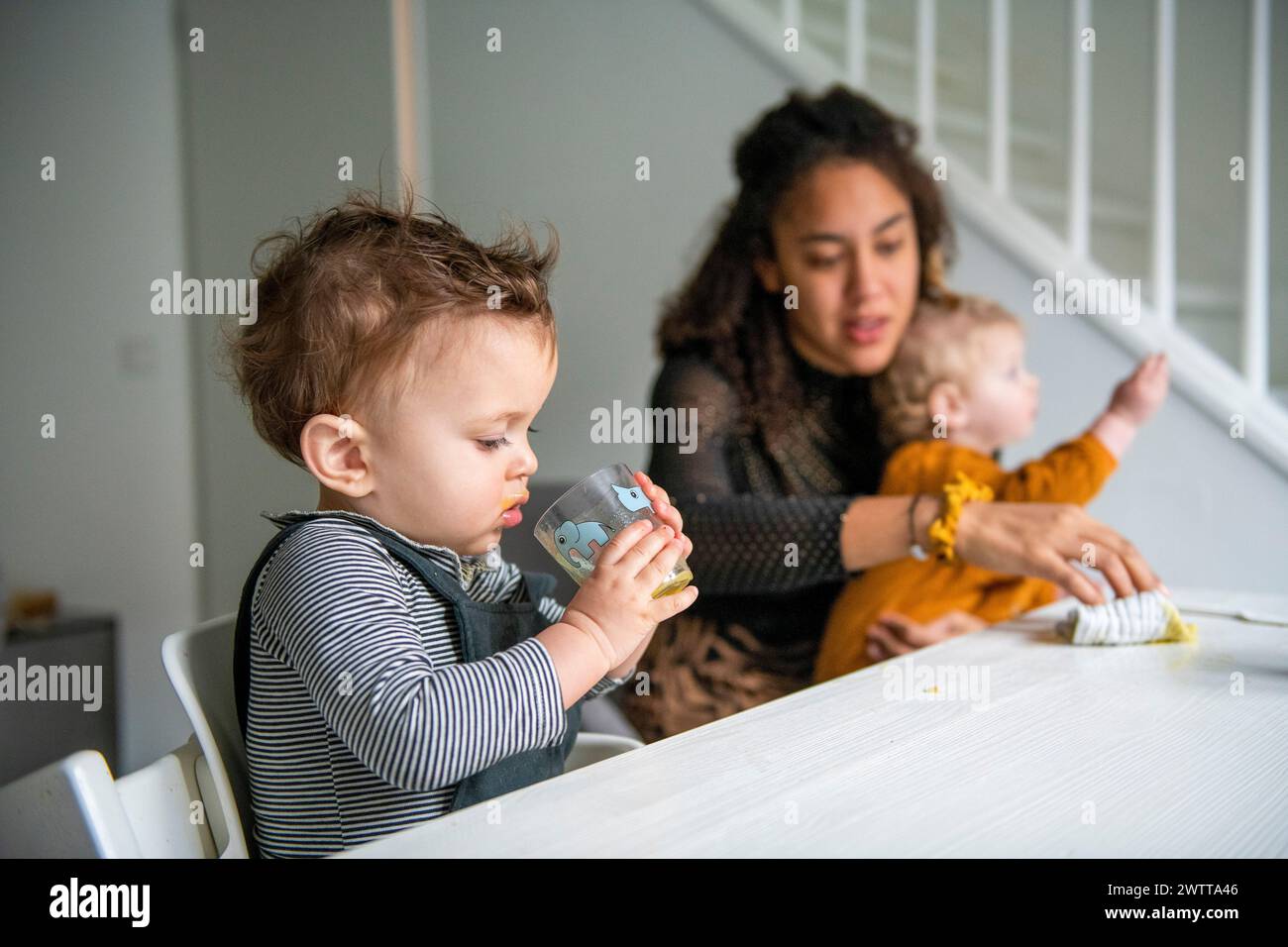Ein kleines Kind schlürft aus einer Tasse, während eine Mutter am Esstisch über zwei Kinder wacht. Stockfoto