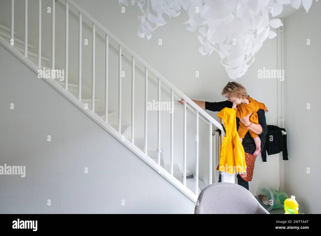 Ein Kind in gelbem Kleid, das die weiße Indoor-Treppe hinuntergeht und sich am Geländer hält, mit einer einzigartigen weißen Deckenbefestigung darüber. Stockfoto