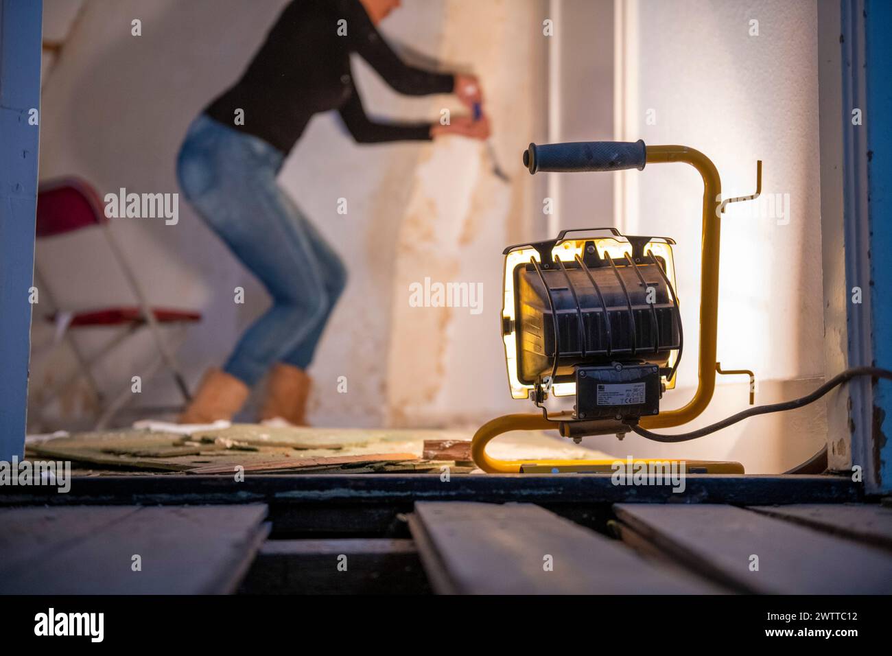 Ein fokussierter Mitarbeiter arbeitet an einer Wandrenovierung, beleuchtet durch eine helle, tragbare Arbeitsleuchte. Stockfoto