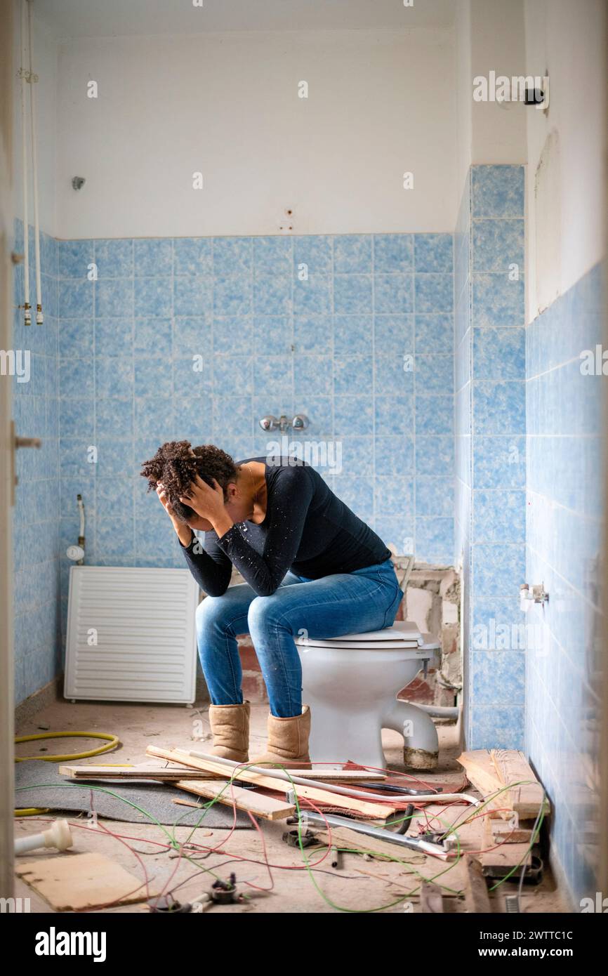 Überwältigte Person, die in einem Raum im Bau auf einer Toilette sitzt, Kopf in Händen. Stockfoto
