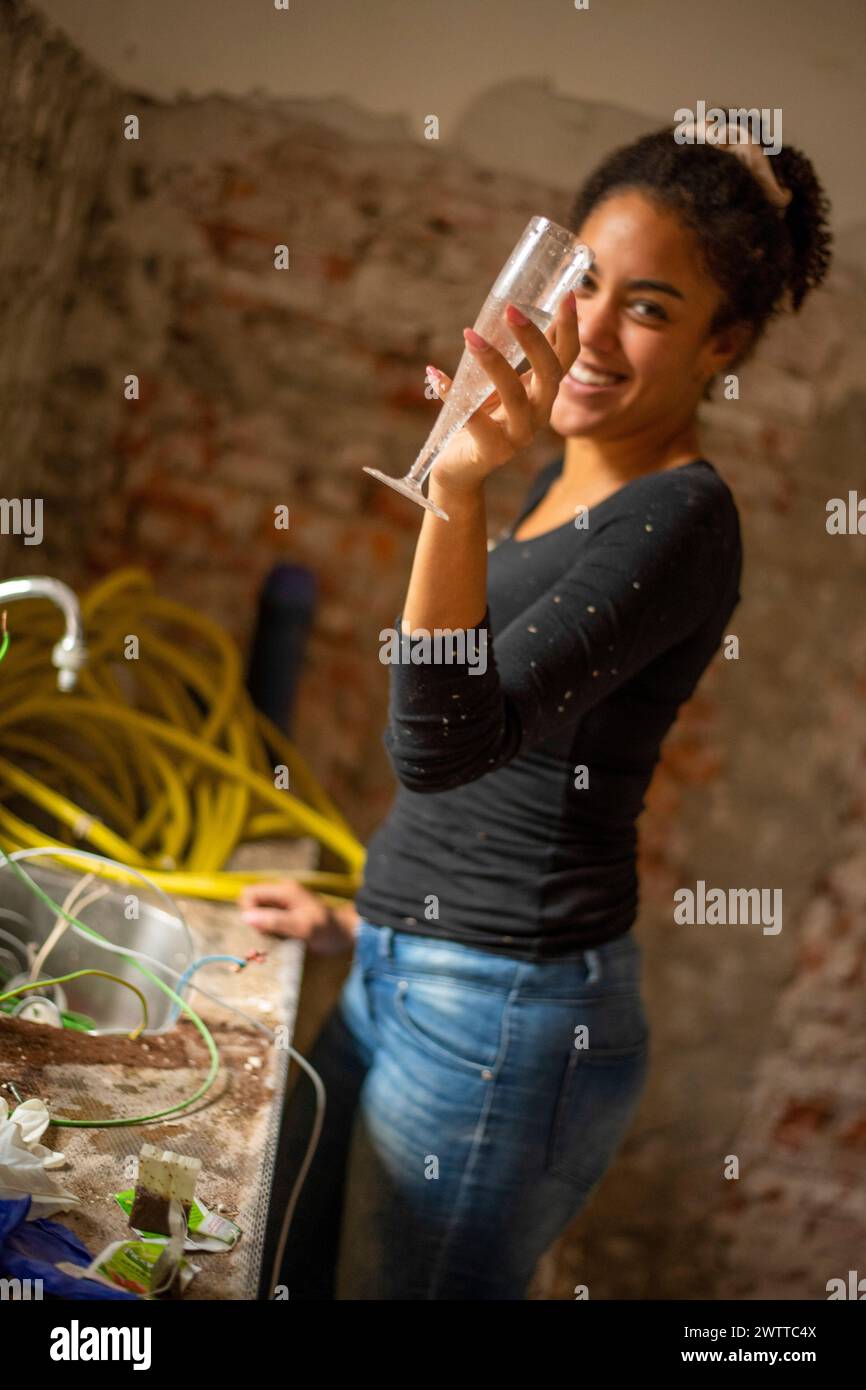 Frau, die mit einem Lächeln in einer schwach beleuchteten Küche im Renovierungschaos tostet. Stockfoto