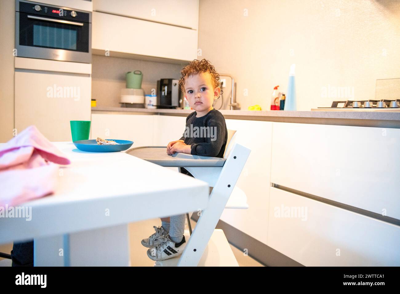 Ein kleines Kleinkind, das mit neugierigem Blick am Küchentisch essen konnte. Stockfoto