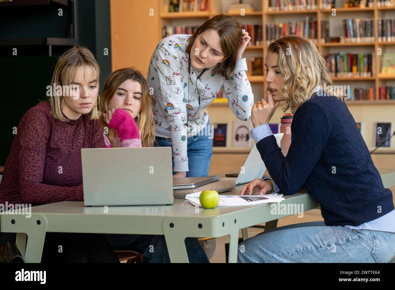 Eine Gruppe von vier jungen Frauen konzentrierte sich auf einen Laptop-Bildschirm, während sie an einem Tisch mit Büchern, Papieren und einem Apfel saßen und schlug eine Studie oder Arbeitssitzung vor. Stockfoto