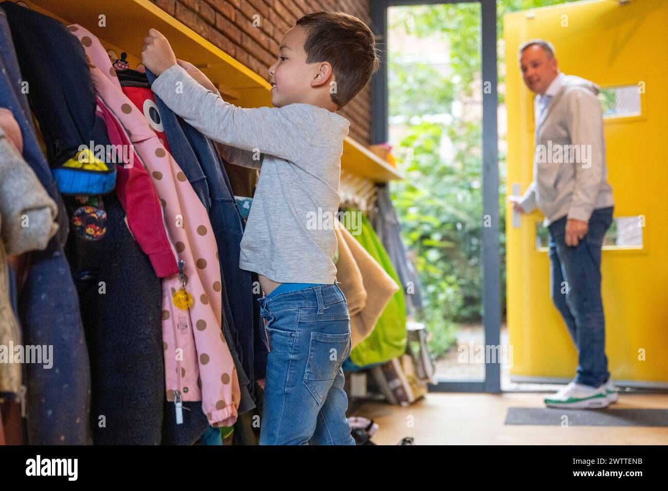 Junge hängt seinen Mantel in der Schule mit einem Lehrer im Hintergrund. Stockfoto