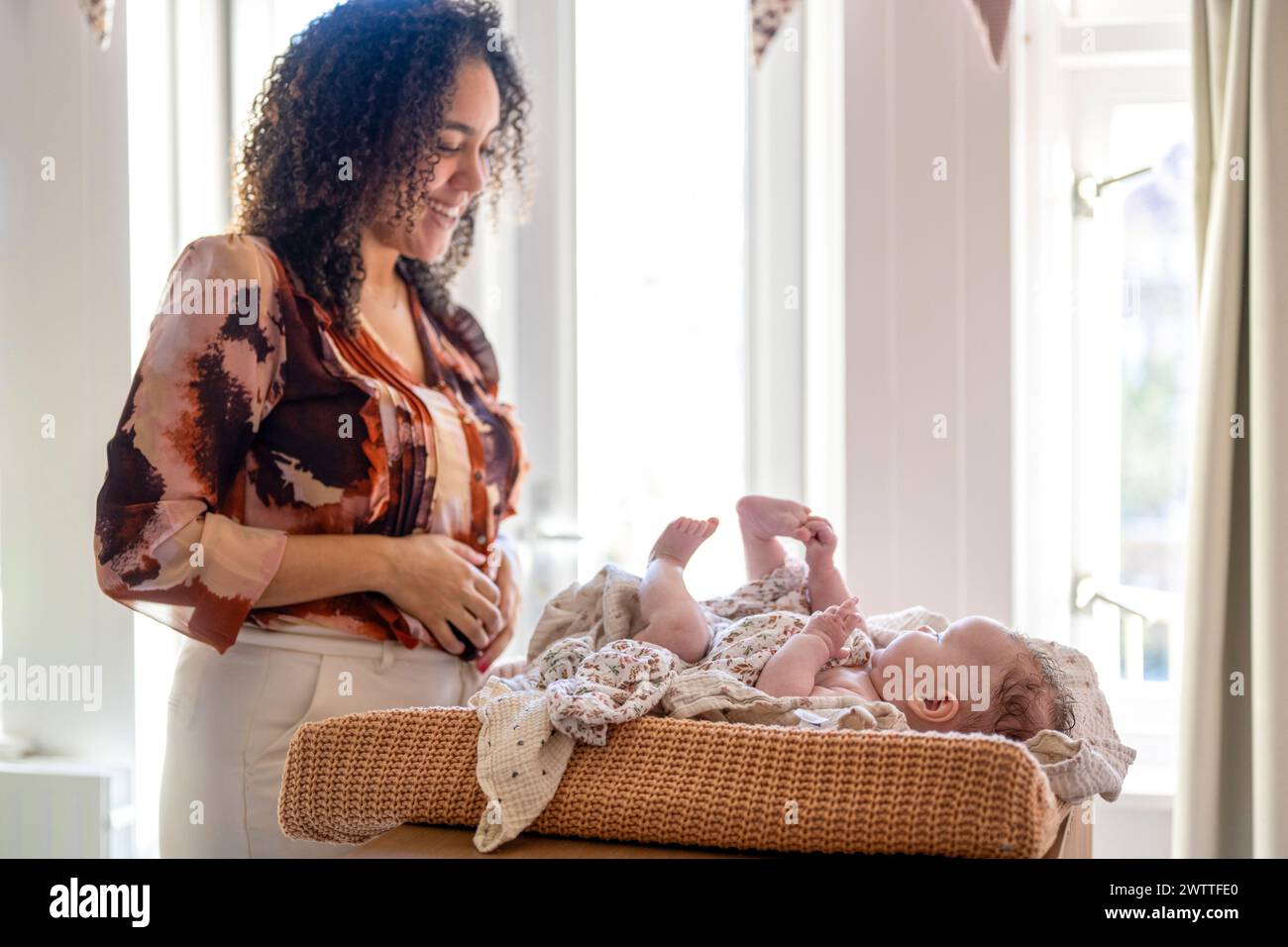 Eine lächelnde Frau, die an einem Fenster steht, berührt sanft ein Baby, das in einer gemütlichen Wanne liegt. Stockfoto