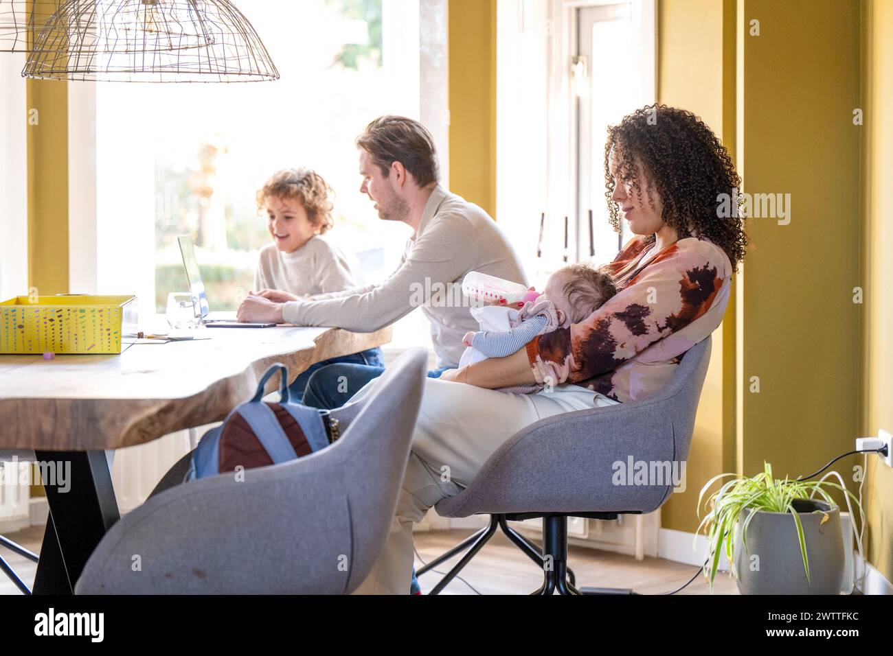 Ein gemütlicher Familienmoment zu Hause mit einer stillenden Mutter und einem Vater, der einem anderen Kind bei den Hausaufgaben hilft. Stockfoto