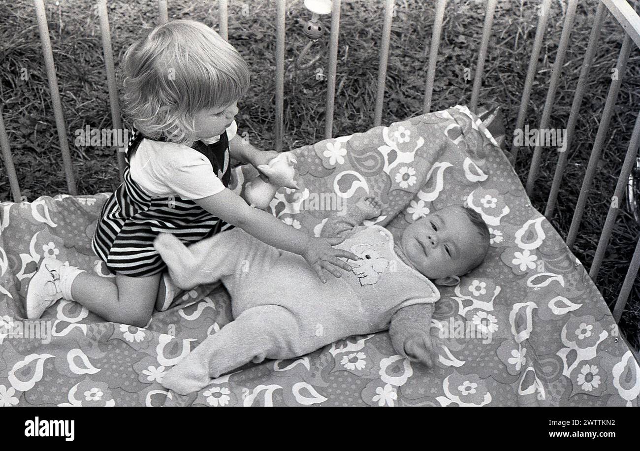 1970er Jahre, historisch, in einem hölzernen Laufstall, ein kleines Mädchen, das mit ihrem kleinen Bruder interagiert, der auf dem Rücken liegt und nach oben blickt, England, Großbritannien. Stockfoto