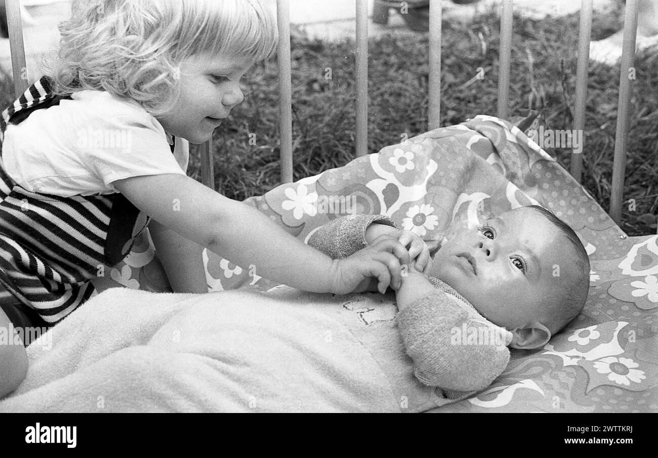 1970er Jahre, historisch, in einem hölzernen Laufstall, ein kleines Mädchen, das mit ihrem kleinen Bruder interagiert, der auf dem Rücken liegt und nach oben blickt, England, Großbritannien. Stockfoto