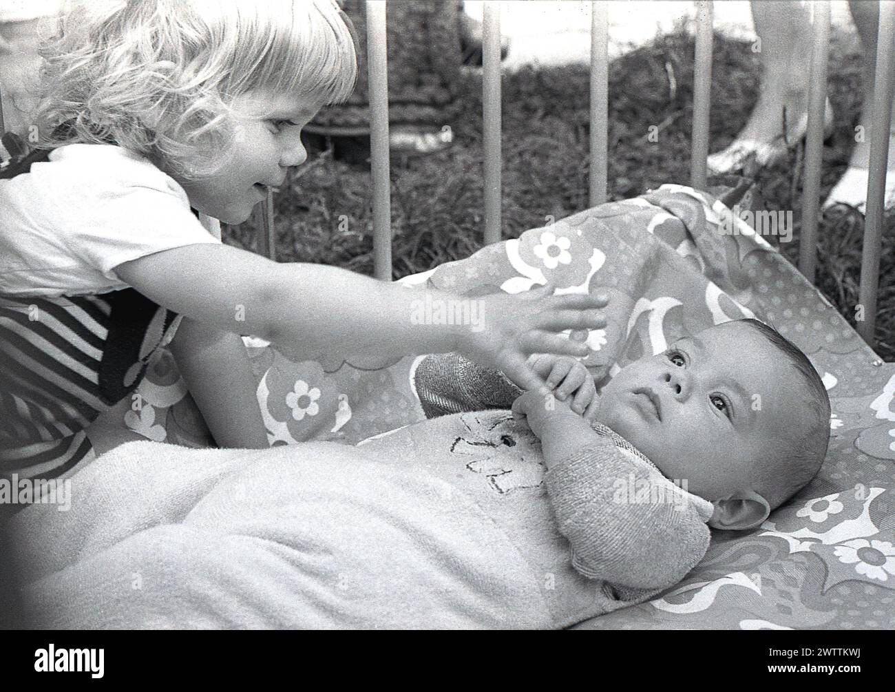 1970er Jahre, historisch, ein kleines Mädchen, das mit ihrem kleinen Bruder interagiert und auf seinem Rücken liegt in einem hölzernen Laufstall, England, Großbritannien. Stockfoto
