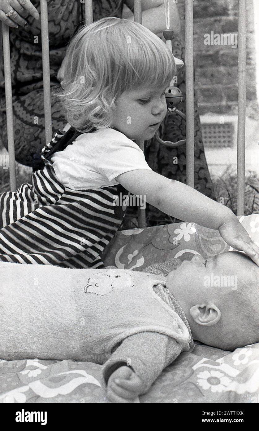1970er Jahre, historisch, ein kleines Mädchen, das mit ihrem kleinen Bruder interagiert und auf seinem Rücken liegt in einem hölzernen Laufstall, England, Großbritannien. Stockfoto