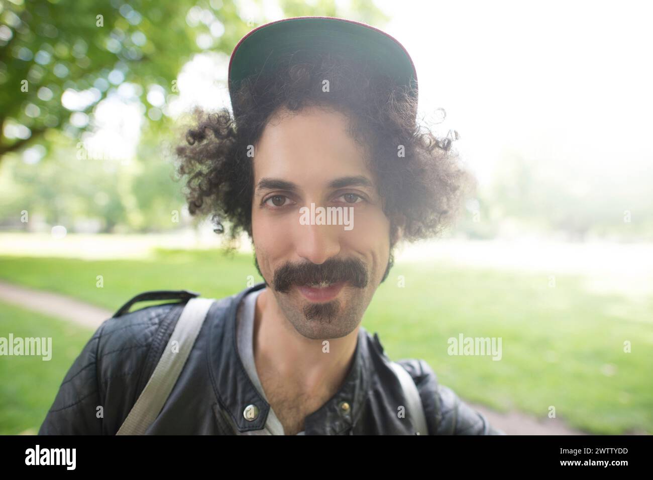 Mann mit lockigen Haaren, der eine Mütze trägt und im Park lächelt Stockfoto